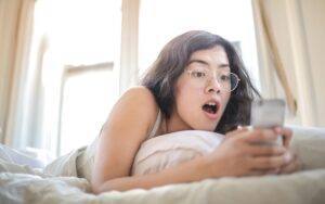 Young woman lying on bed, surprised by something on her smartphone, in a bright bedroom setting.